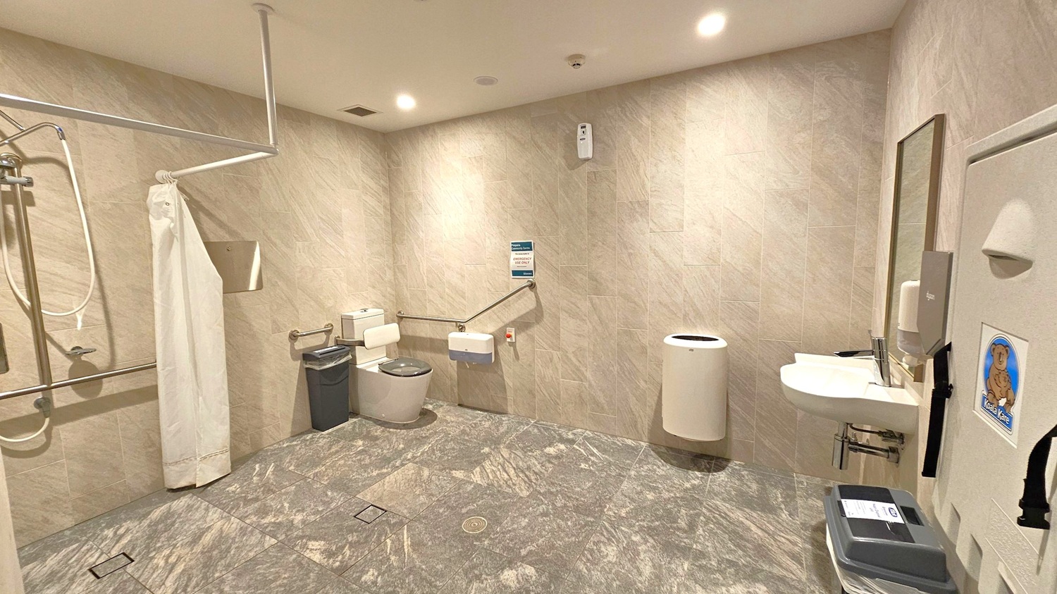 A spacious, accessible bathroom with light grey tiled walls and floor. On the left, a shower area with a white curtain and grab bars. In the centre, a toilet with support rails and a small bin nearby. On the right, a sink with a mirror above it, a soap dispenser, and a baby changing station with a koala logo. The ceiling has recessed lighting, and a white cylindrical bin is positioned against the wall.