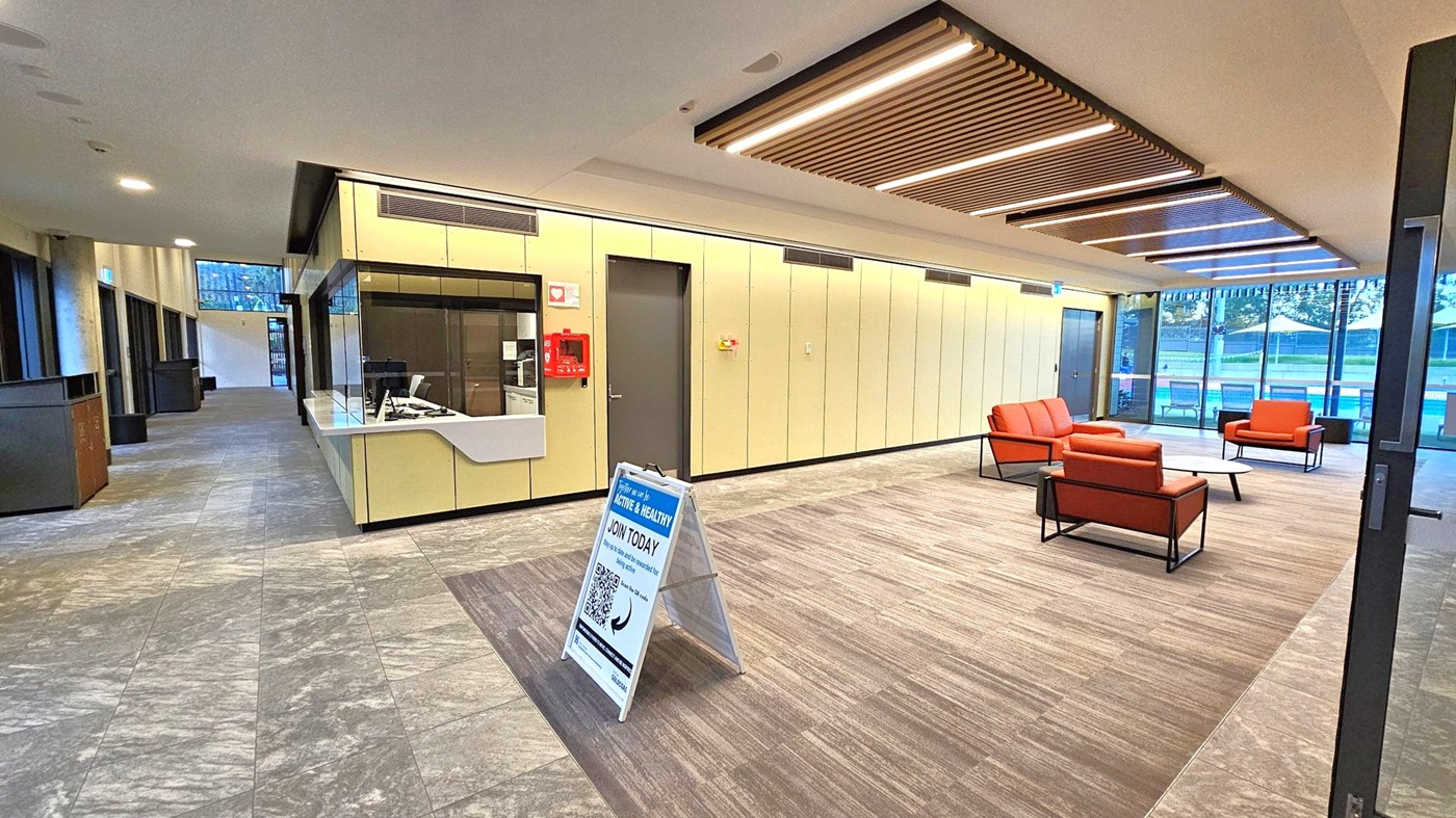 A modern interior space with a reception desk on the left, featuring a computer and a fire extinguisher on the wall. In the centre, a signboard with information about joining a programme. On the right, a seating area with orange chairs and a table, set on a carpeted floor. Large windows in the background reveal an outdoor area with tables and umbrellas. The ceiling has recessed lighting and wooden slats, while the floor is a mix of tile and carpet.