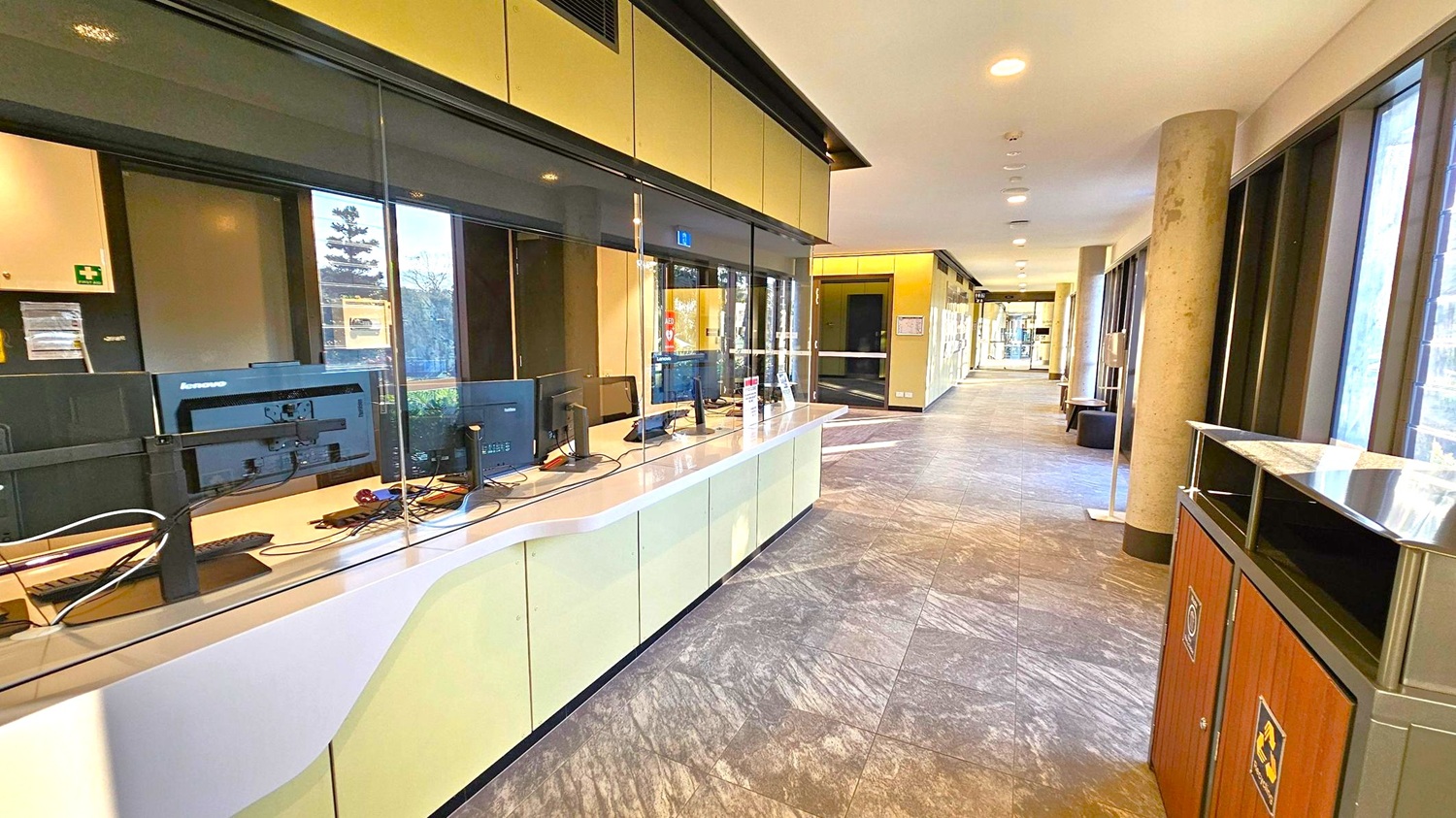 A modern office reception area with a long, curved desk on the left, equipped with multiple computer monitors and a glass partition. The floor is covered with grey tiles, and the ceiling has recessed lighting. On the right, large windows allow natural light to enter, and there are cylindrical columns along the corridor. A recycling bin is visible in the foreground on the right. The corridor extends into the distance, with additional seating and doors visible.