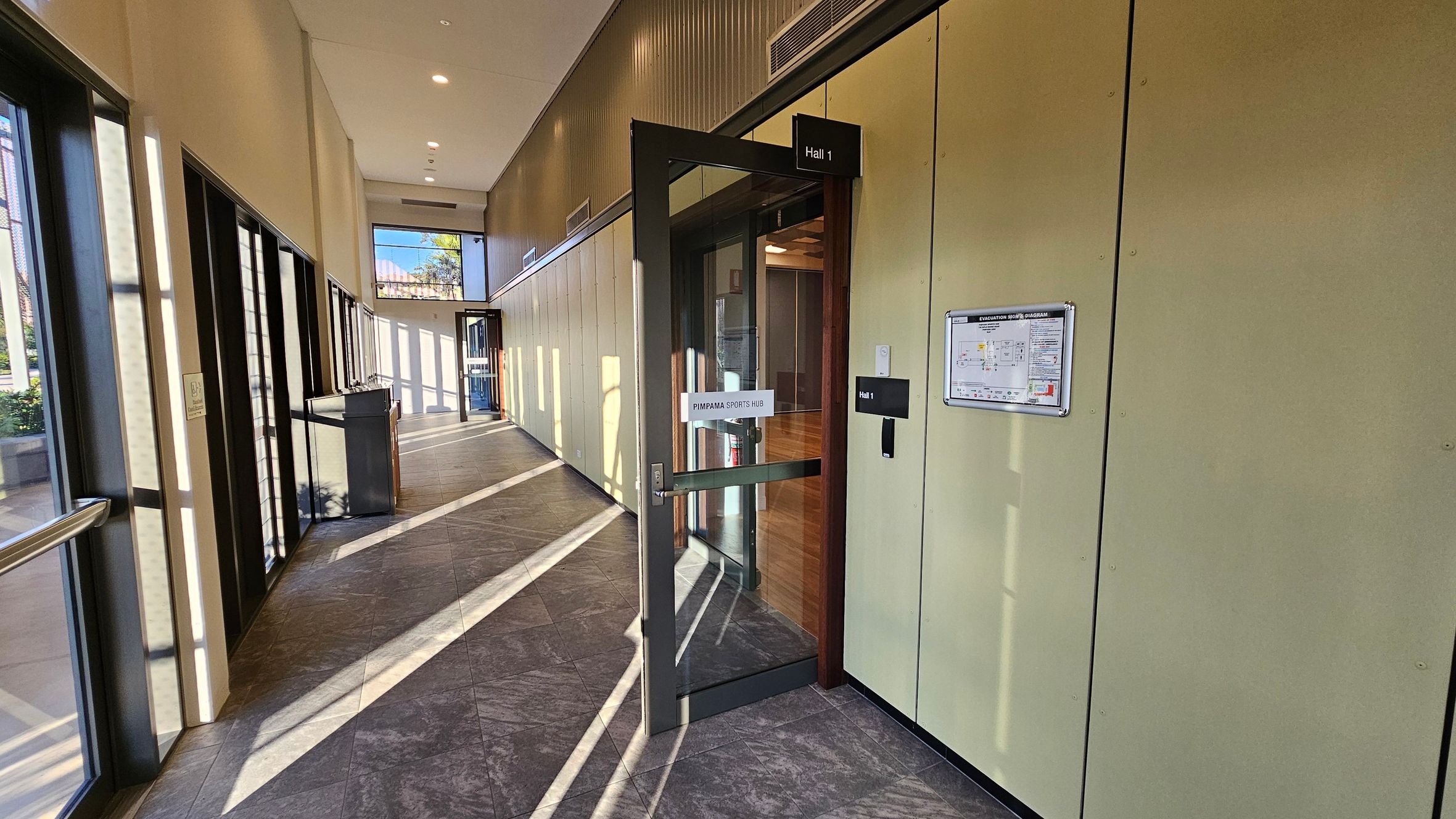 A corridor with large windows on the left, allowing sunlight to cast shadows on the tiled floor. On the right, there is a door labelled "Hall 1" with a sign reading "Pimpama Sports Hub." Next to the door is an evacuation route diagram. The ceiling is high with recessed lighting, and the walls are light-coloured. At the end of the corridor, there is another door leading outside, with visible greenery through the glass.