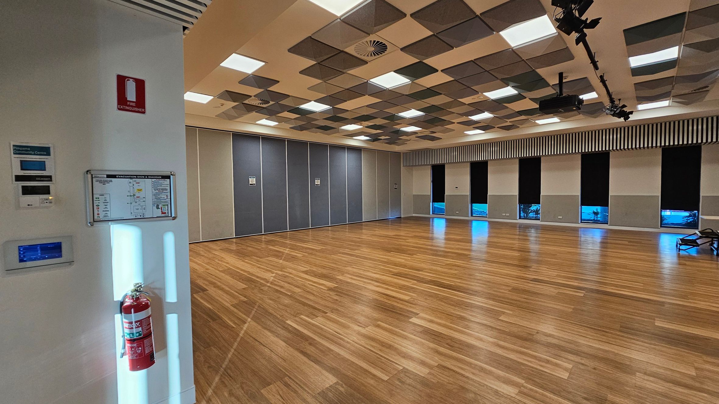 A spacious room with a polished wooden floor. On the left, a white wall features a fire extinguisher, an evacuation sign, and control panels. The ceiling has a pattern of square panels with integrated lights and ventilation. The right side has large windows with black blinds, allowing some light through. The back wall is lined with grey partition panels. A few chairs are stacked in the far right corner.