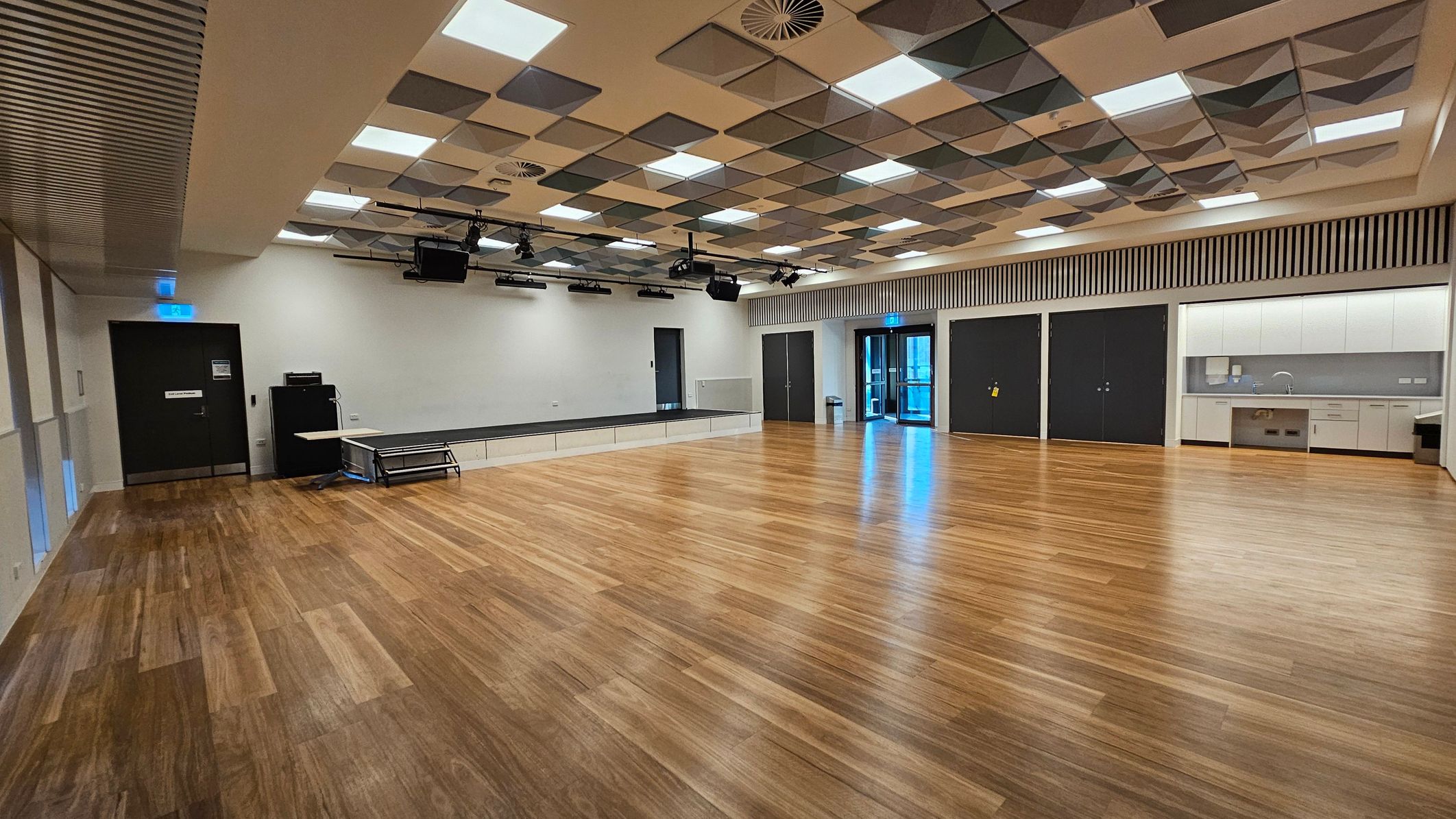 A spacious room with a wooden floor and a ceiling featuring a pattern of square panels in various shades. On the left, there is a black door with an exit sign above it, and a stack of tables and equipment nearby. The right side has a kitchenette area with white cabinets and a sink. In the centre, there are large black double doors and a glass door leading outside. The room is well-lit with ceiling lights and has a modern, minimalist design.