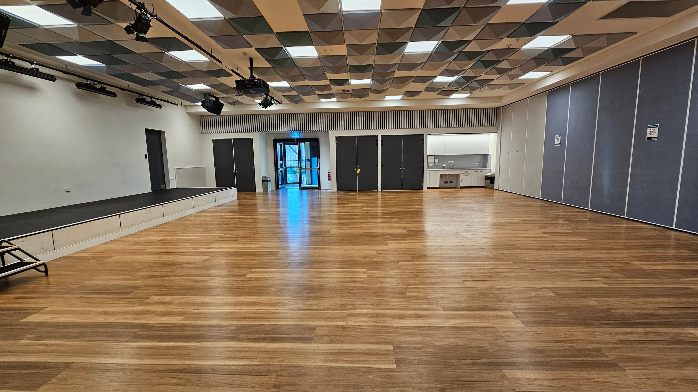 A spacious room with a wooden floor and a patterned ceiling featuring square panels. On the left, there's a small stage with black steps leading up to it. In the centre, double doors with glass panels are visible, flanked by dark doors. On the right, a kitchenette area with cabinets and a sink is present, along with large, grey partition walls. The ceiling has a mix of recessed lights and mounted black speakers or lighting equipment.