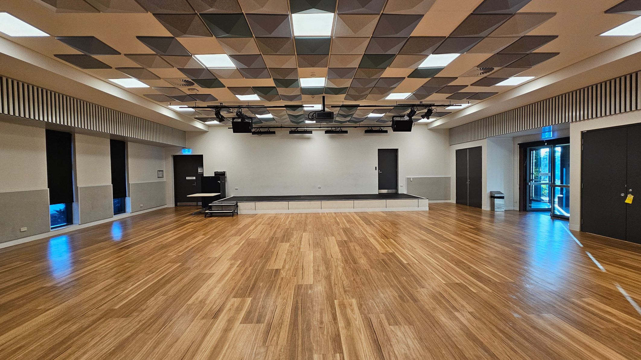 A spacious room with a wooden floor and a patterned ceiling featuring square panels. In the centre, there is a low stage with a black surface. On the left, there are several tall windows with black coverings. On the right, a set of double doors with glass panels leads outside. The walls are light-coloured with a striped design near the ceiling. Lighting fixtures and speakers are mounted on the ceiling above the stage.