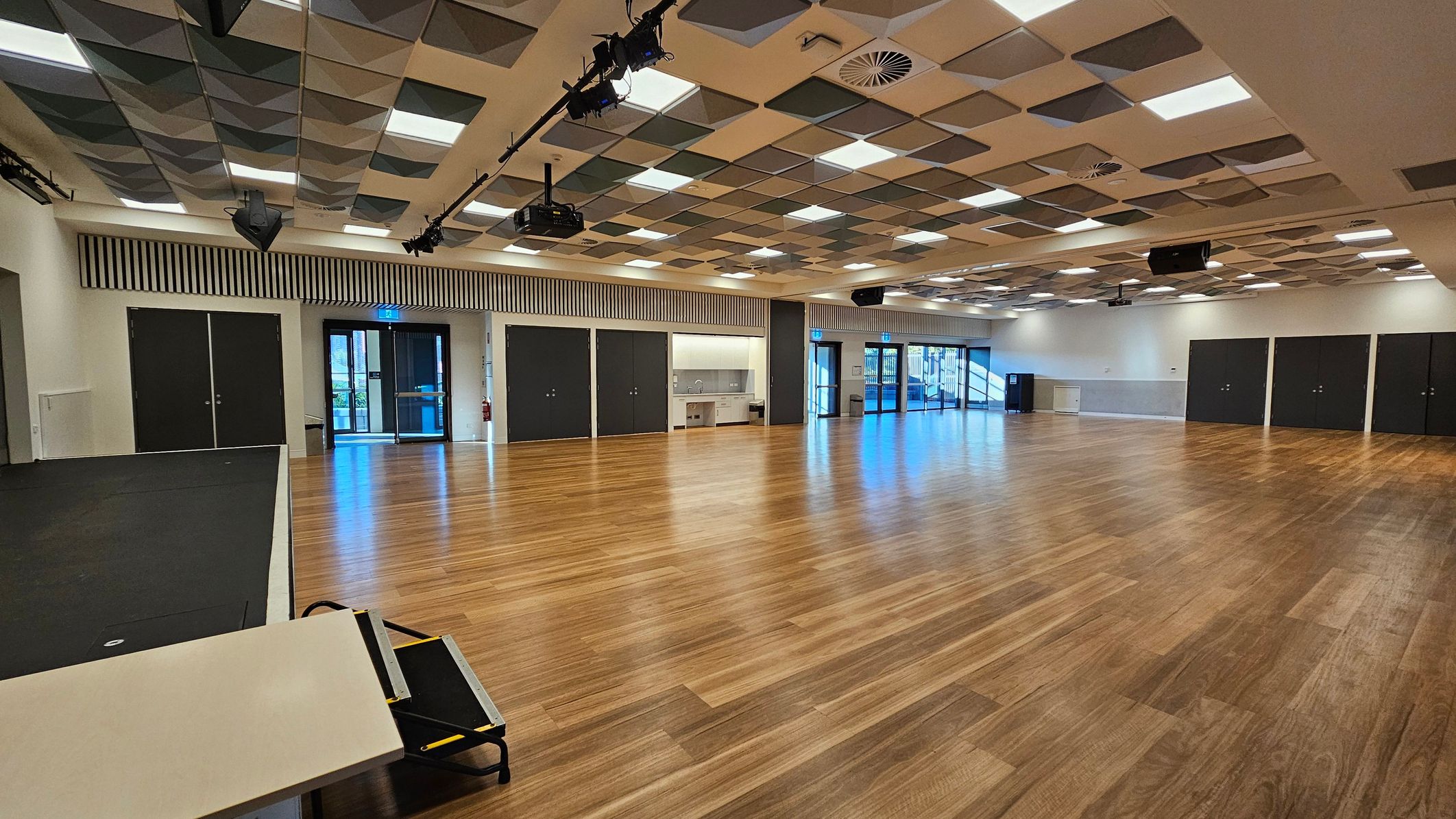 A spacious room with a wooden floor and a patterned ceiling featuring square panels in various shades. On the left, there is a small stage area with a black surface and a table with a folded chair. The back wall has several black double doors and a kitchenette area with a sink. Large glass doors on the right lead outside. The ceiling has lighting fixtures and speakers, and the room is well-lit with natural light from the glass doors.