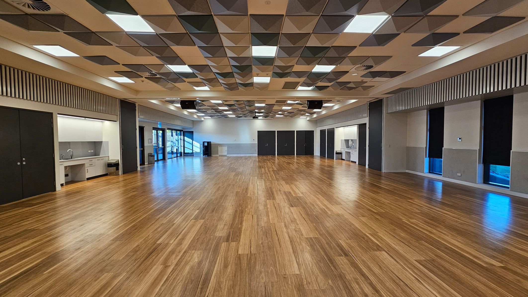 A spacious room with a polished wooden floor. The ceiling features a geometric pattern of squares and triangles with integrated lighting. On the left, there is a kitchenette area with a sink and cabinets. The right side has large windows with black blinds. The back wall has several dark doors and a small counter. The room is well-lit, with natural light coming through the windows.