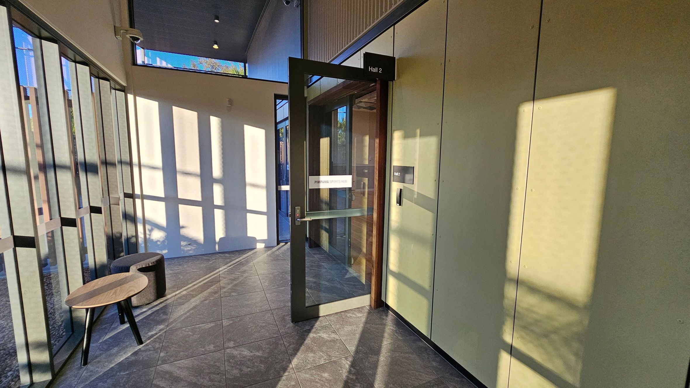 A hallway with a glass door labelled "Hall 2" on the right, leading into a room. The door has a sign reading "Pimpama Sports Hub". The left side features large windows casting shadows on the tiled floor. A small wooden table and a round, cushioned stool are placed near the windows. The ceiling is high with visible light fixtures.