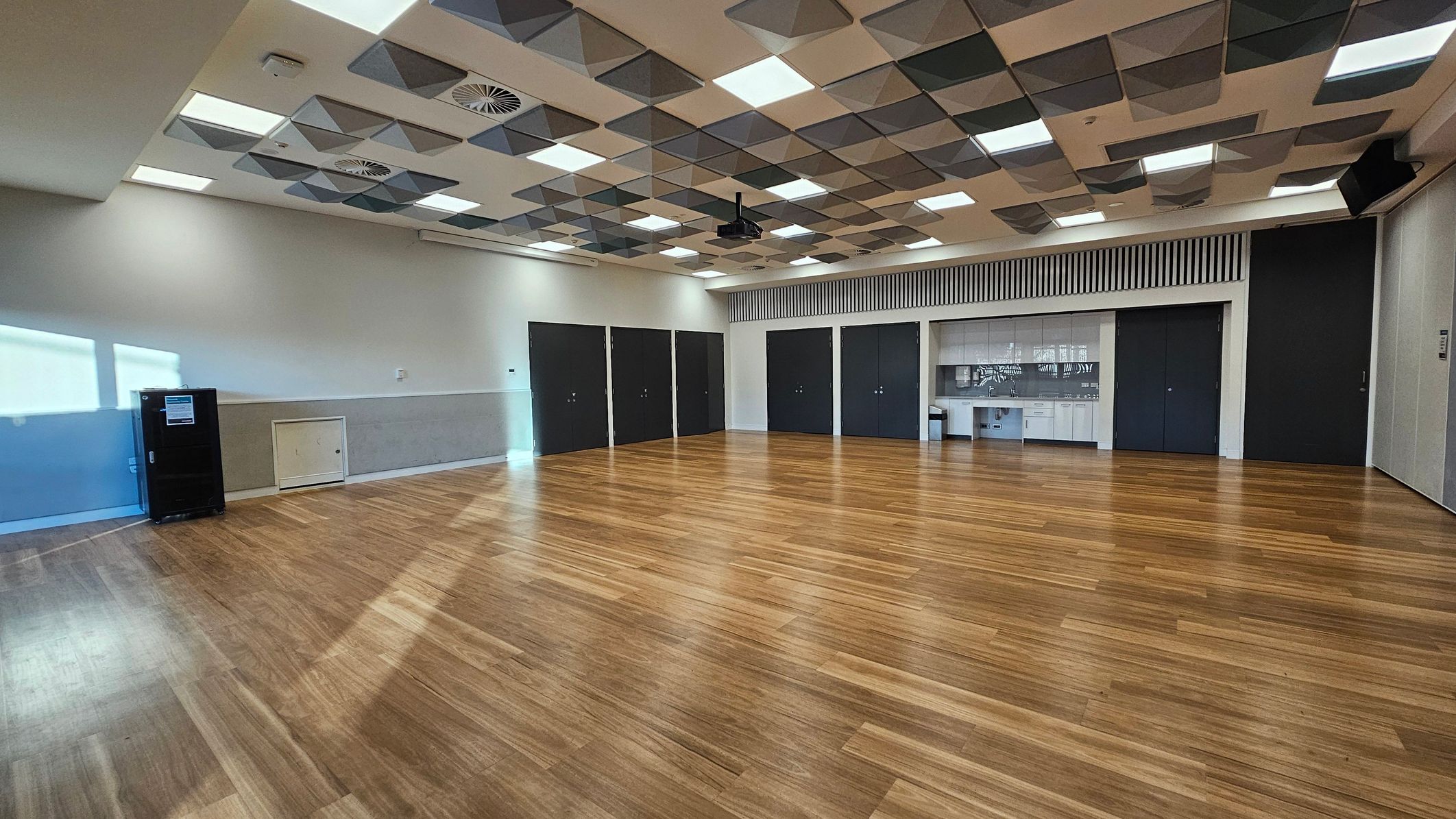 A spacious room with a wooden floor and a ceiling featuring a geometric pattern of recessed lights and ventilation panels. On the left, there is a black cabinet against a light-coloured wall. The back wall has several dark doors and a section with a kitchenette, including a sink and cupboards. The room is well-lit with natural light coming from windows on the left.