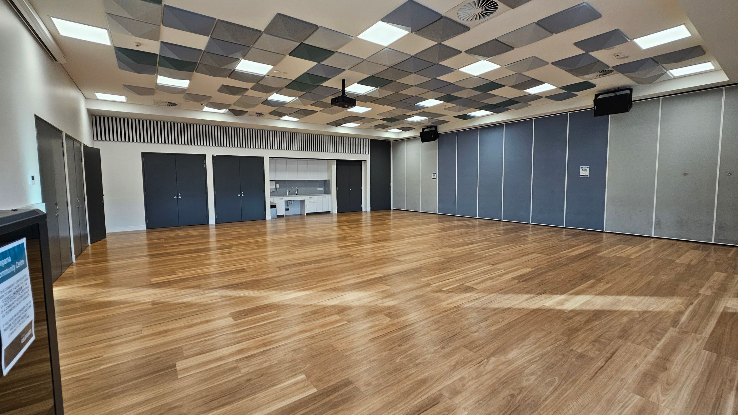 A spacious room with a wooden floor and a ceiling featuring a pattern of square panels in various shades. On the left, there are dark double doors and a notice board. In the centre, a kitchenette with a sink and cabinets is visible, flanked by more dark doors. The right side has large, blue-grey partition panels. The room is well-lit with multiple ceiling lights.