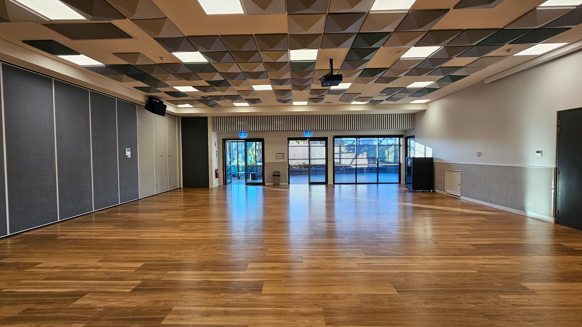 A spacious room with a wooden floor and a ceiling featuring a geometric pattern of recessed panels and lights. On the left, there are large grey partition walls. The back wall has three sets of glass doors leading outside, with a small bin and a fire extinguisher nearby. On the right, a dark door and a small cabinet are visible. The room is well-lit, with natural light streaming in through the glass doors.