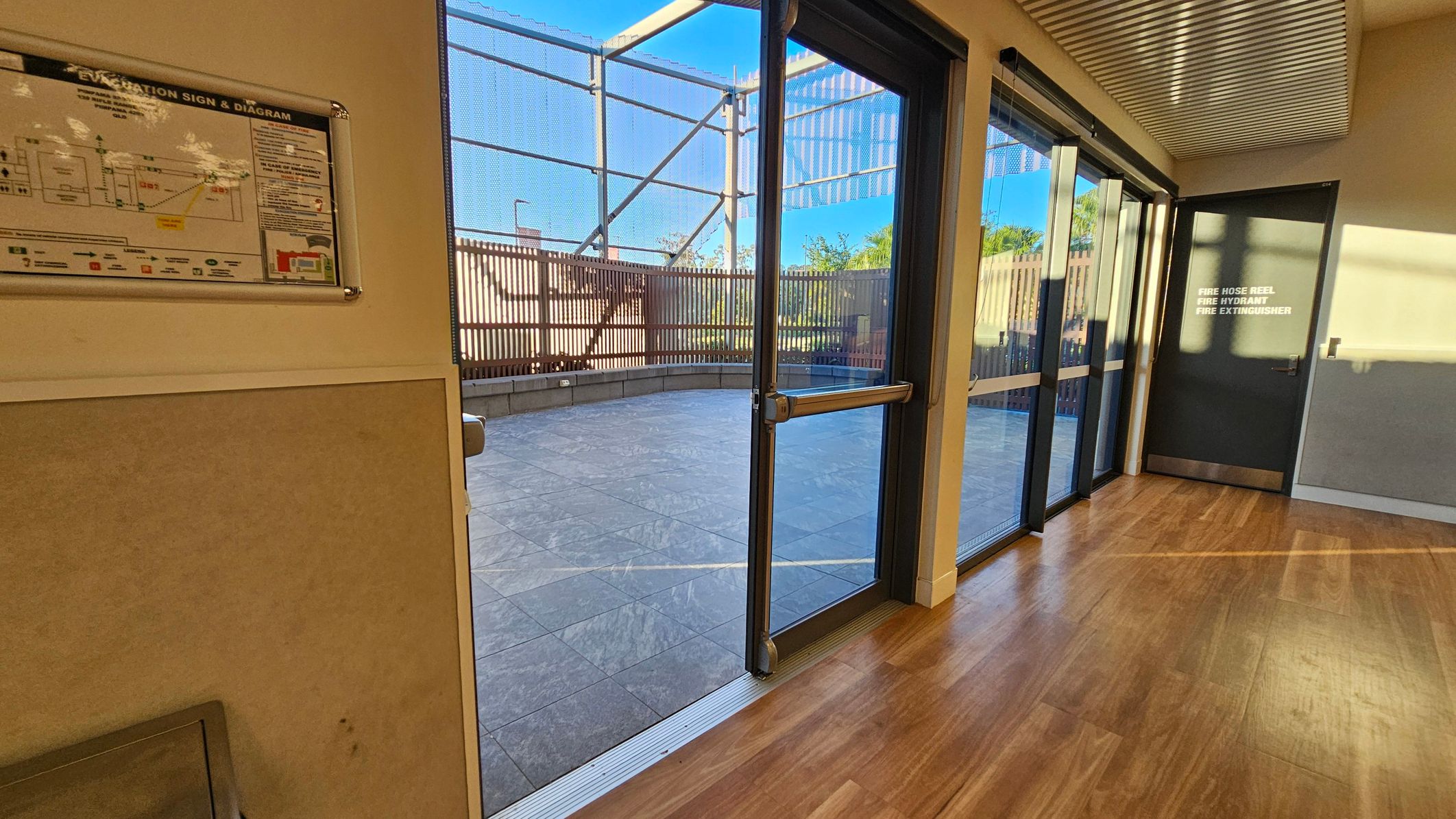 A corridor with wooden flooring and a metal-panelled ceiling. On the left, there's an evacuation sign and diagram on the wall. In the centre, large glass doors open to an outdoor area with a tiled floor and a metal fence. On the right, a door marked for fire safety equipment is visible. Sunlight casts shadows on the floor and walls, creating a bright atmosphere.