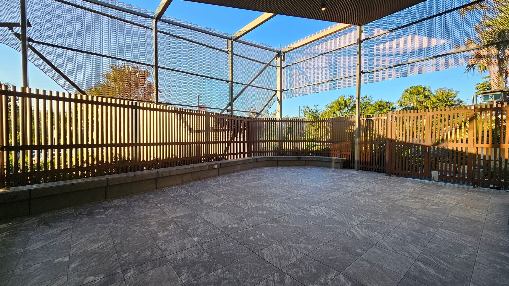 A spacious outdoor area with a grey tiled floor. The space is enclosed by a wooden slat fence and metal mesh panels, allowing visibility of the surrounding greenery. In the background, there are trees and plants visible through the fence. The ceiling is partially covered with a slatted roof, casting shadows on the ground. The area appears to be well-lit with natural light.