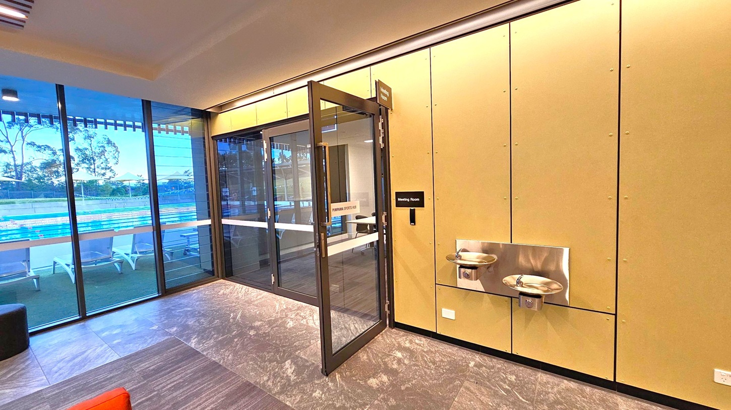 On the left, large glass windows reveal an outdoor swimming pool with lanes and sun loungers. The middle features a glass door leading to a meeting room, with a sign above and on the door. On the right, a beige wall has two stainless steel water fountains. The floor is covered with grey and brown tiles, and the ceiling has recessed lighting.