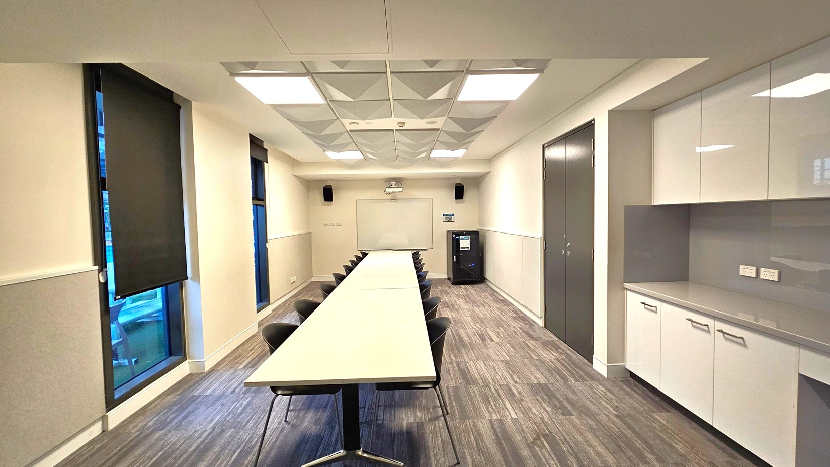 A modern conference room with a long rectangular table in the centre, surrounded by black chairs. On the left, there are tall windows with black blinds. The right side features a wall with white cabinets and a countertop. At the far end, a whiteboard is mounted on the wall with a projector above it. The floor is covered with grey wood-patterned tiles, and the ceiling has recessed lighting and geometric panels.