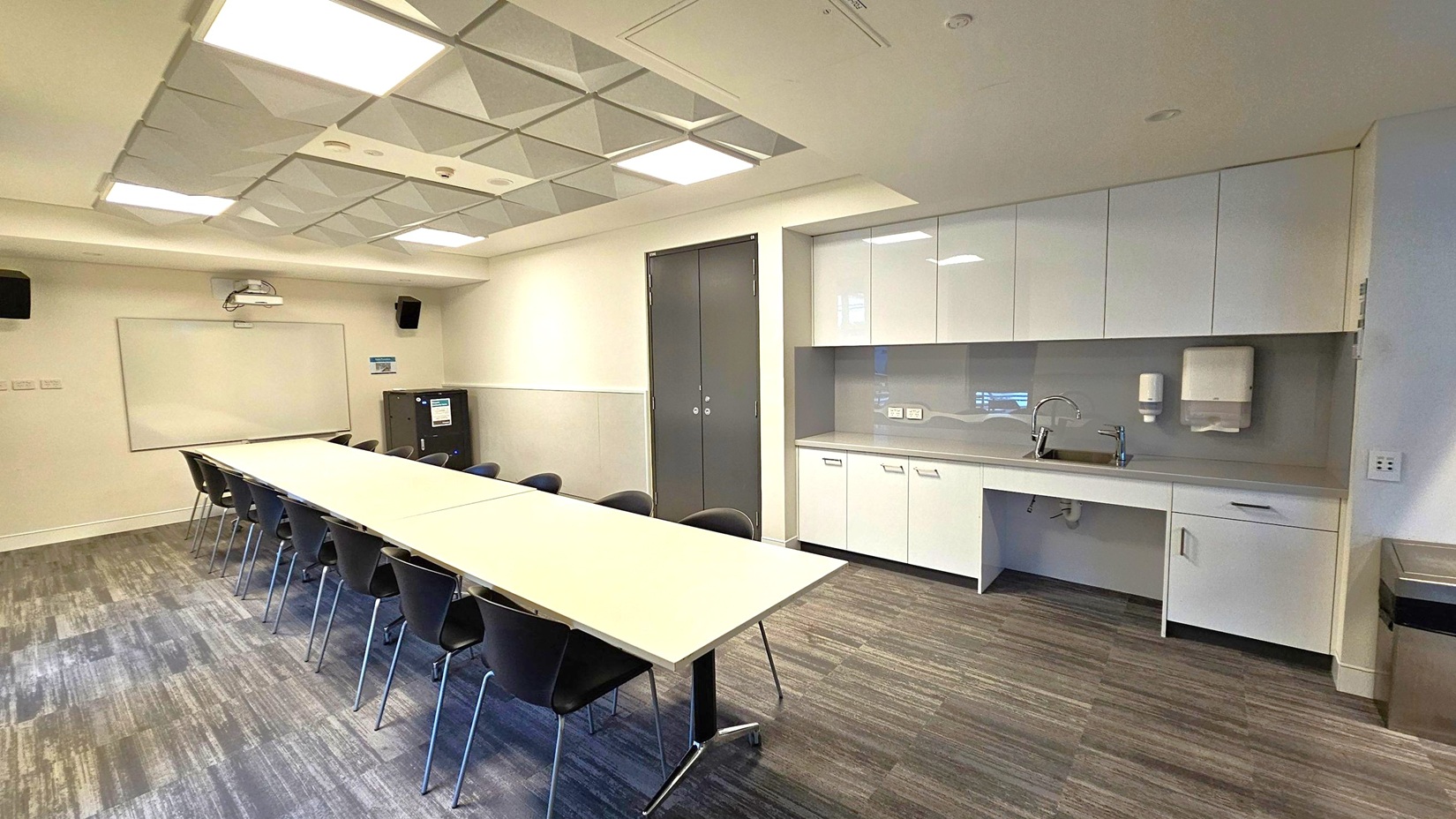 A modern conference room with a long rectangular table in the centre, surrounded by black chairs. On the right, there is a kitchenette with white cabinets, a sink, and a paper towel dispenser. The back wall features a whiteboard and a projector. The ceiling has recessed lighting and a grid pattern. The floor is carpeted with a mix of grey and green tones.