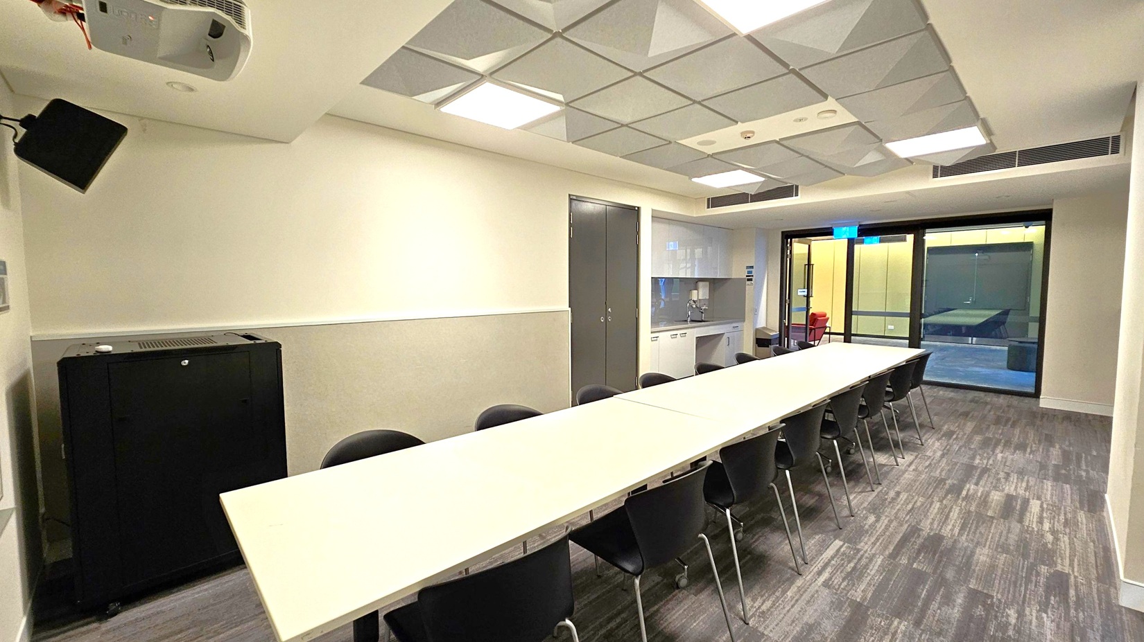 A conference room with a long white table in the centre, surrounded by black chairs. On the left, there's a black cabinet and a wall-mounted speaker. The ceiling has square recessed lights and a grid pattern. On the right, there's a kitchenette with a sink and cabinets, and a glass door leading to another room. The floor is carpeted with a grey and green pattern.