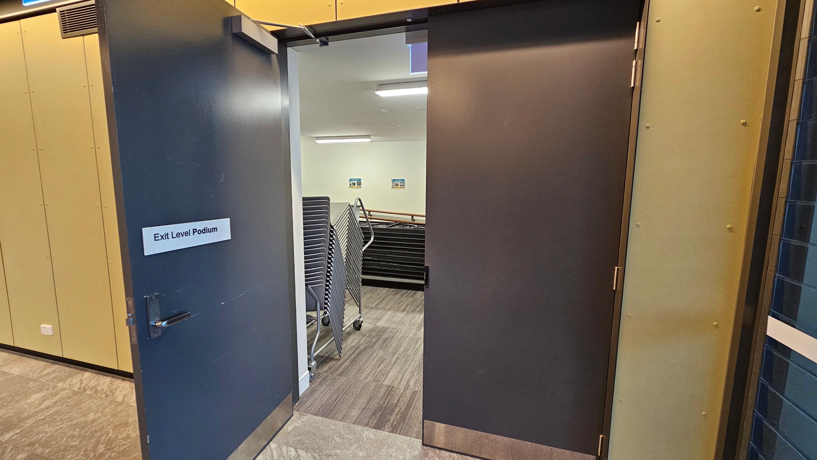 A dark grey door is partially open, revealing a room with a stack of chairs on trolleys. The door has a sign reading "Exit Level Podium". The floor is covered with light brown tiles. Inside, the walls are white with two small signs visible. The ceiling has fluorescent lights. The door frame and adjacent wall are a light beige colour.