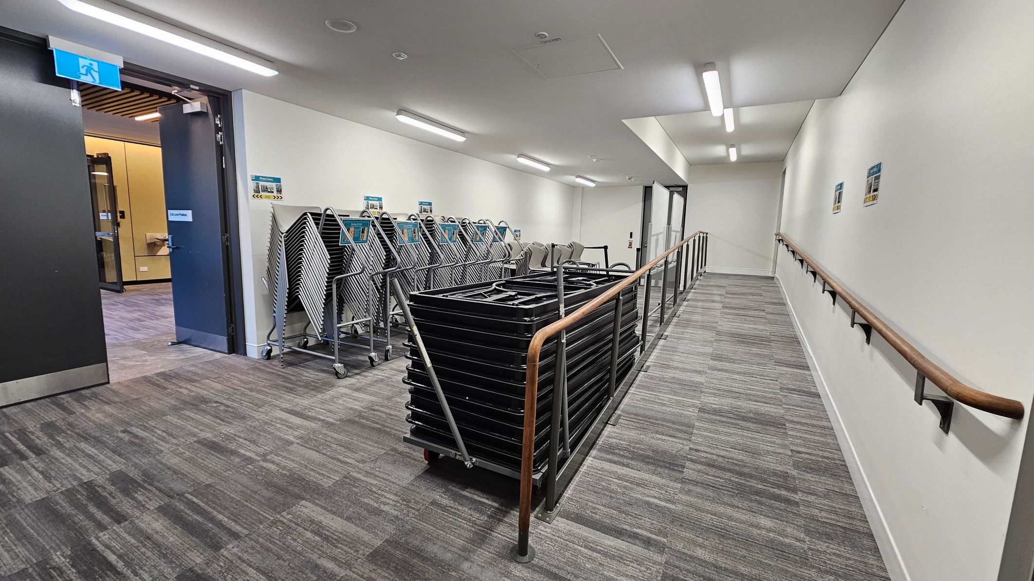 A corridor with grey carpeted flooring and white walls. On the left, an open door leads to a room with a wooden floor and a blue exit sign above. Near the door, several stacks of folded chairs are on trolleys. On the right, a handrail runs along the wall, and there are posters attached to the wall. The ceiling has fluorescent lights evenly spaced along the corridor.