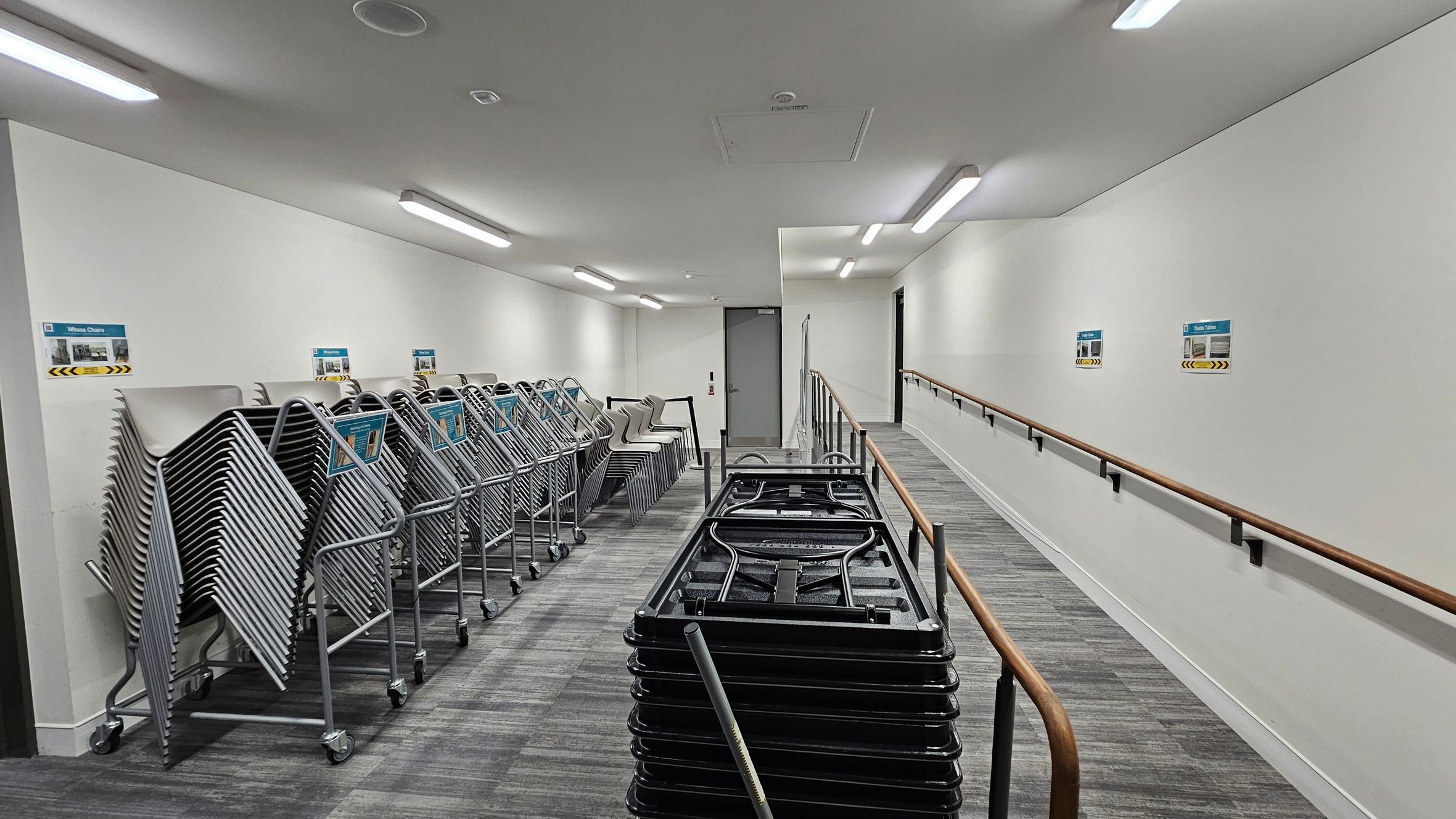 A corridor with a grey floor and white walls. On the left, there are stacks of metal folding chairs on trolleys. On the right, a wooden handrail runs along the wall, with several signs above it. In the centre, a stack of folded tables is visible. The ceiling has several rectangular fluorescent lights. At the end of the corridor, there is a closed door.