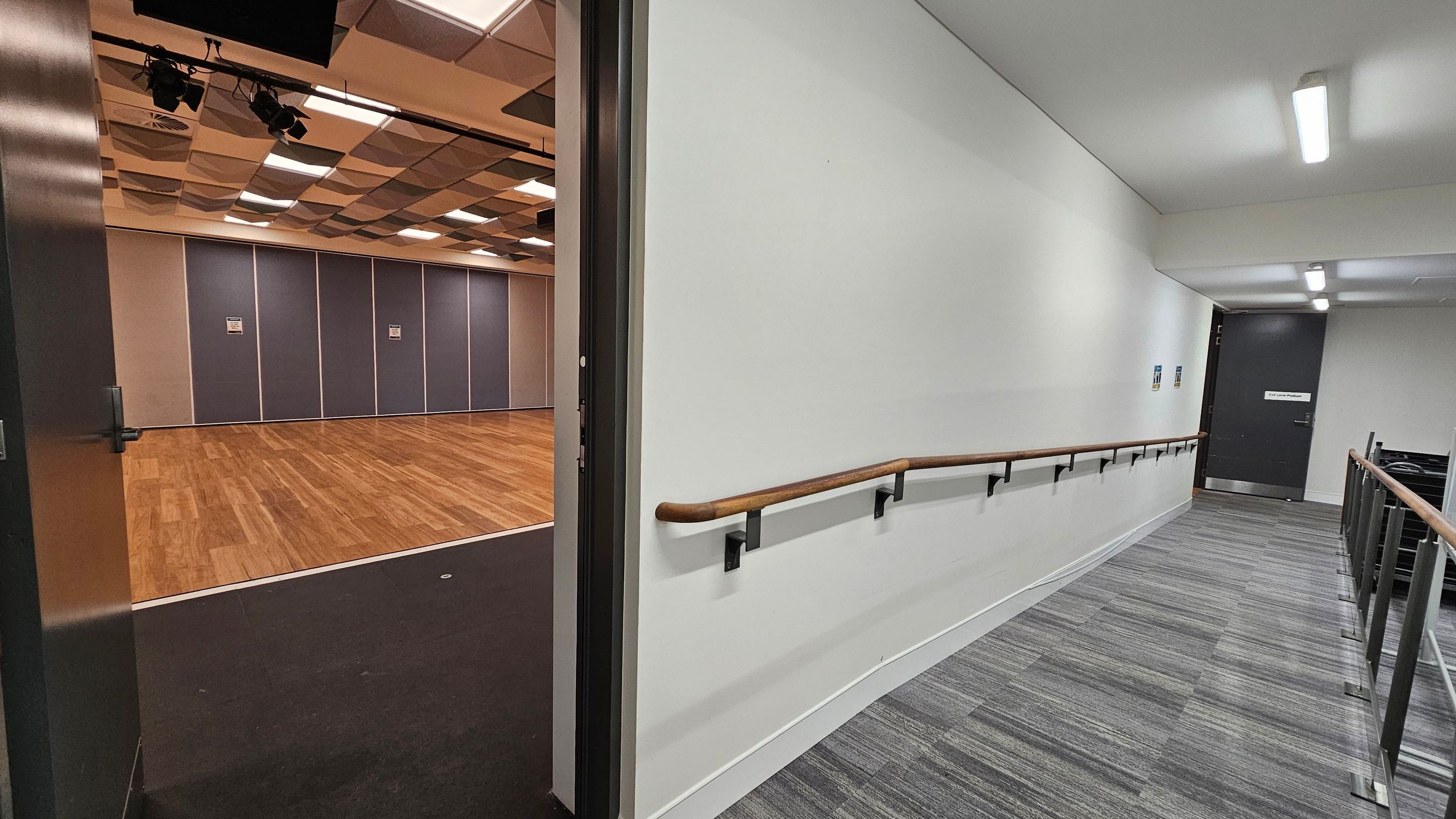 A corridor with a grey carpeted floor and white walls, featuring a wooden handrail on the right side. The ceiling has fluorescent lights. On the left, an open door leads to a room with a wooden floor and purple wall panels. The room's ceiling has a pattern of square acoustic panels and several stage lights.