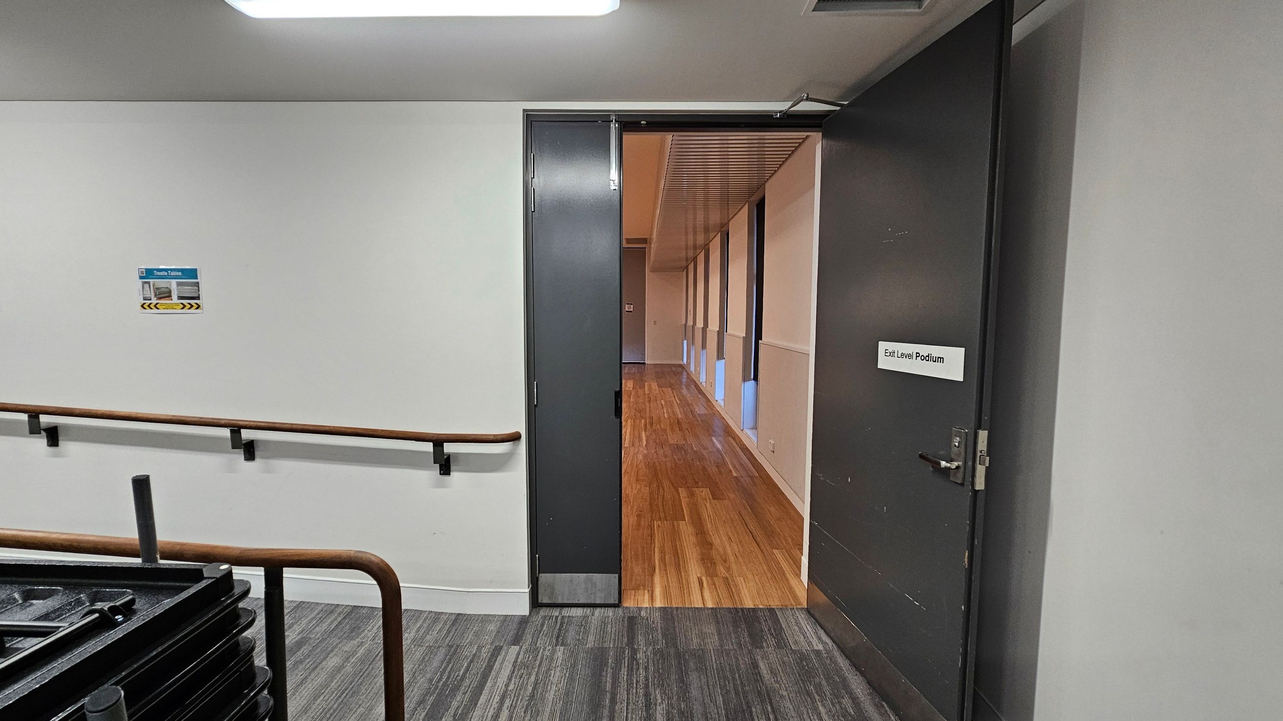 A hallway with a grey door on the right, slightly open, revealing a corridor with wooden flooring and a series of windows on the right. The door has a sign reading "Exit Level Podium." On the left, a white wall features a sign above a wooden handrail. The floor in the foreground is covered with grey carpet tiles, and a stack of black tables is partially visible. The ceiling has a fluorescent light fixture.