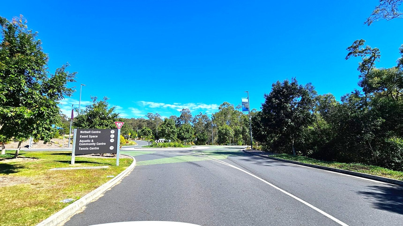 A road curves to the right, bordered by lush green trees on both sides. On the left, a sign lists directions to various centres, including a netball centre and aquatic centre. The road features a roundabout with green markings. Street lamps and a banner are visible on the right. The area is surrounded by dense foliage, creating a natural setting.