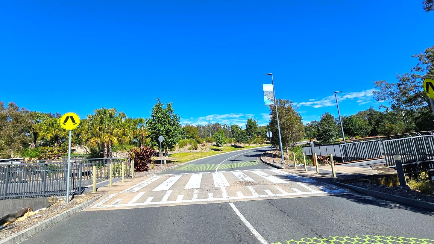 A pedestrian crossing with a yellow warning sign on the left. The road curves to the right, flanked by greenery and trees. A metal railing runs along the right side, with a footpath beside it. A banner hangs from a streetlight on the right. The ground is paved, and the crossing has a striped pattern.
