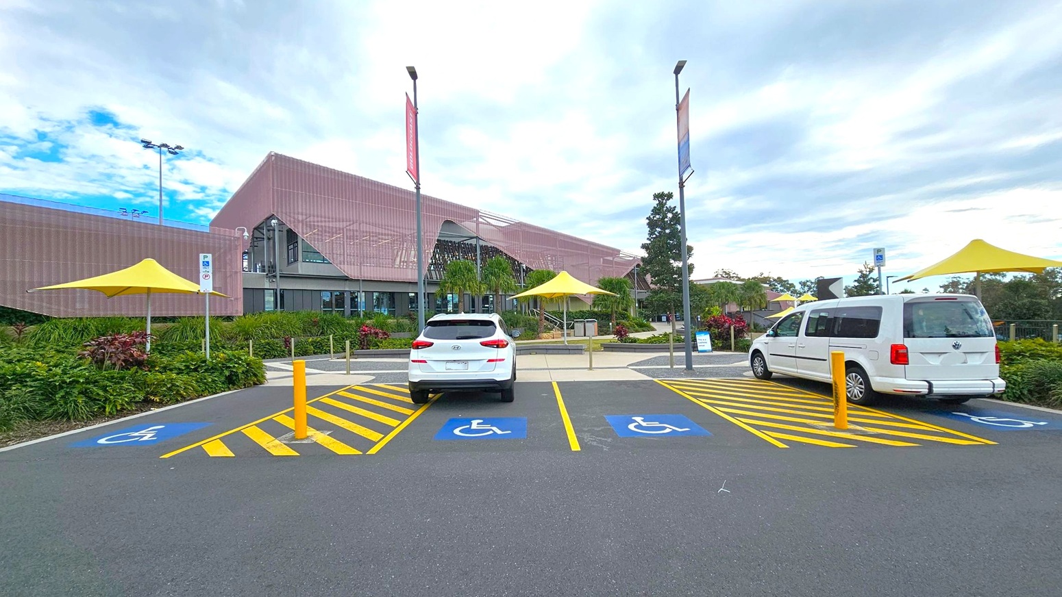 A modern building with a pinkish facade is in the background, featuring large glass windows and surrounded by greenery. In the foreground, there are two parked white vehicles in designated disabled parking spaces, marked with blue and yellow lines. Yellow umbrellas provide shade over nearby seating areas. The ground is paved, and tall streetlights are visible.
