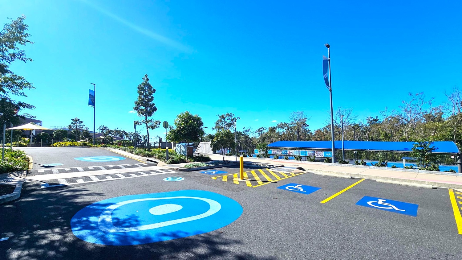 A parking area with designated disabled parking spaces marked in blue on the asphalt. On the right, there are two spaces with wheelchair symbols. A yellow-striped area separates them. In the background, a blue-roofed structure runs parallel to the parking spaces. On the left, a pedestrian crossing leads to a path lined with greenery and trees. Tall streetlights and banners are visible along the path. The area is surrounded by trees and shrubs.