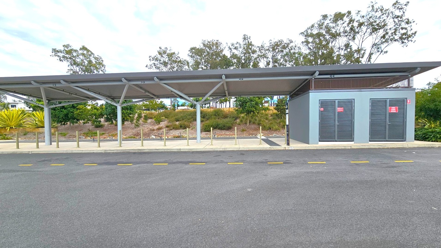 A bus stop with a modern shelter featuring a slanted roof supported by metal beams. On the right, there's a grey utility building with two louvred doors displaying red signs. The foreground has a paved area with yellow dashed lines on the road. In the background, there are trees and shrubs on a slightly elevated grassy area. The overall setting is clean and organised.