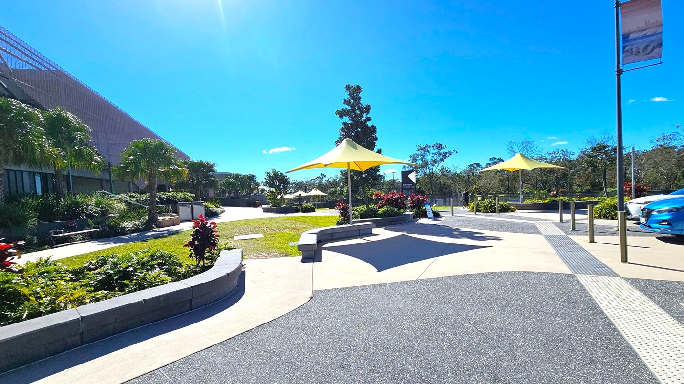 A paved outdoor area with yellow triangular shade structures. On the left, there are lush green plants and palm trees alongside a large building with a textured facade. In the centre, a pathway leads through landscaped gardens with more plants and trees. On the right, a blue car is parked near metal bollards. The ground is a mix of textured and smooth surfaces, and there are signs and banners on poles.