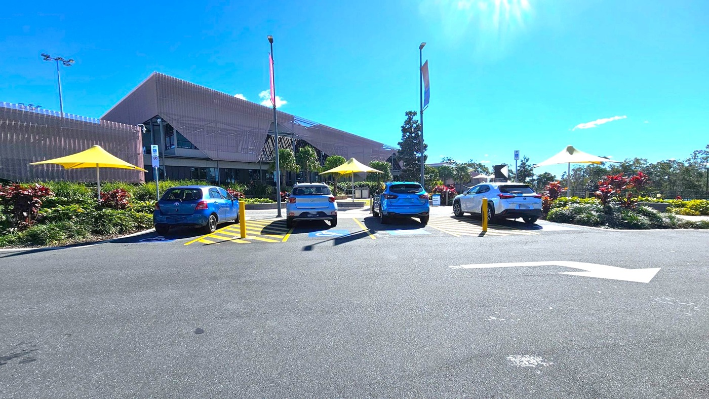 A car park with four vehicles in designated disabled parking spaces, marked by blue signs and yellow lines. Two yellow shade structures are visible, one on the left and one in the centre. The background features a large building with a modern design and a garden area with shrubs and trees. A clear blue sky is above, and the asphalt surface of the car park is in the foreground, with a white directional arrow painted on it.