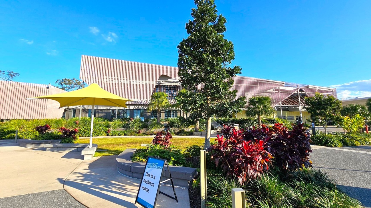 A modern building with a unique, angular design is in the background, featuring a facade with vertical lines. In the foreground, there is a landscaped area with various plants and a prominent tree. A yellow umbrella provides shade over a seating area. A sign reads "This is a cashless venue." The ground is a mix of concrete and gravel paths, and the area is well-maintained with lush greenery.