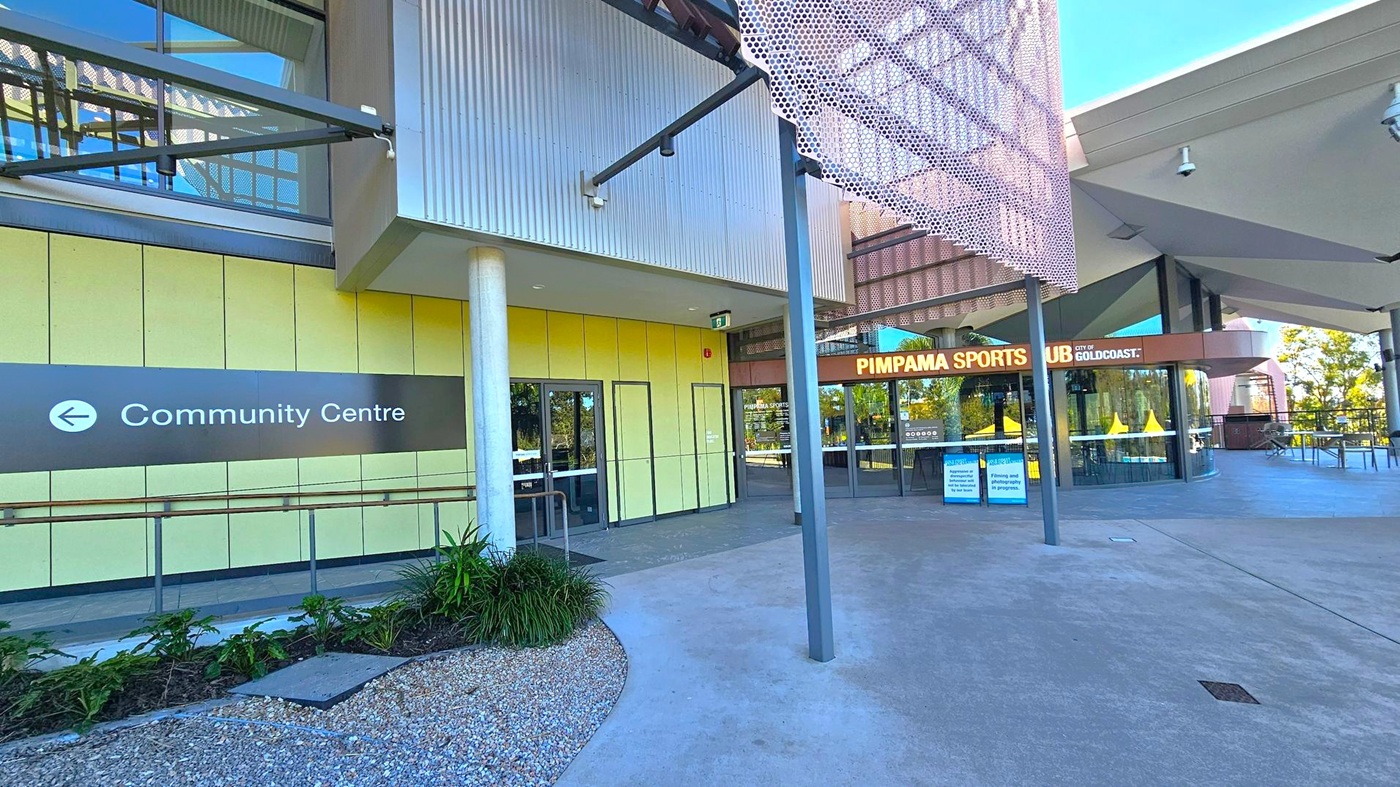 On the left, a sign points to the Community Centre with a pathway leading to it. The building has yellow panels and a glass entrance. In the centre, a column supports the structure. On the right, the entrance to Pimpama Sports Club is visible, with large glass windows and a sign above. The foreground features a gravel area with plants, and the ceiling has a modern design with exposed beams.