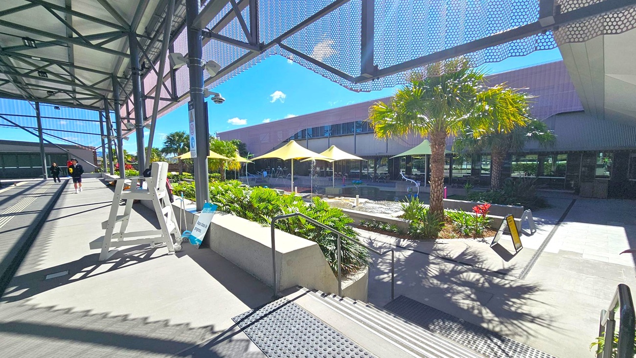 A covered walkway with a metal framework on the left, featuring a white lifeguard chair and a sign. Steps lead down to a courtyard with lush greenery and palm trees. Yellow shade sails cover a seating area in the middle. A modern building with large windows is in the background. The ground is paved, and there are a few people walking in the distance.