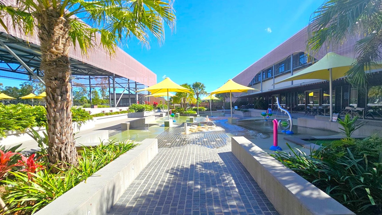 A courtyard with a paved walkway in the centre, flanked by low concrete walls and lush greenery. Yellow umbrellas provide shade over seating areas on both sides. On the left, a large structure with a metal framework is visible, while on the right, a building with large windows and a shaded patio area can be seen. Palm trees and other tropical plants are scattered throughout, and a small water play area with fountains is in the middle.