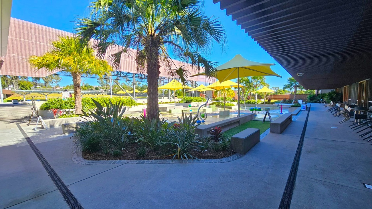 A courtyard with a concrete floor and a row of chairs along the right side under a shaded area. In the centre, there are several yellow umbrellas providing shade over benches and a small water feature. Lush green plants and palm trees are scattered throughout. A large, perforated metal structure is visible in the background, with more greenery and trees beyond it. The sky is clear and blue.