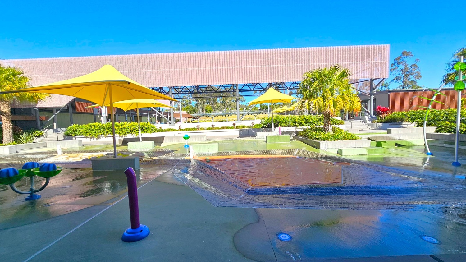 A water play area with a wet surface and various water features, including a purple pipe and blue and green structures. Yellow umbrellas provide shade, and there are green plants and palm trees around. In the background, a large structure with a slatted roof and metal framework is visible, along with more greenery and a staircase leading to an elevated area. The ground is wet, indicating recent water activity.