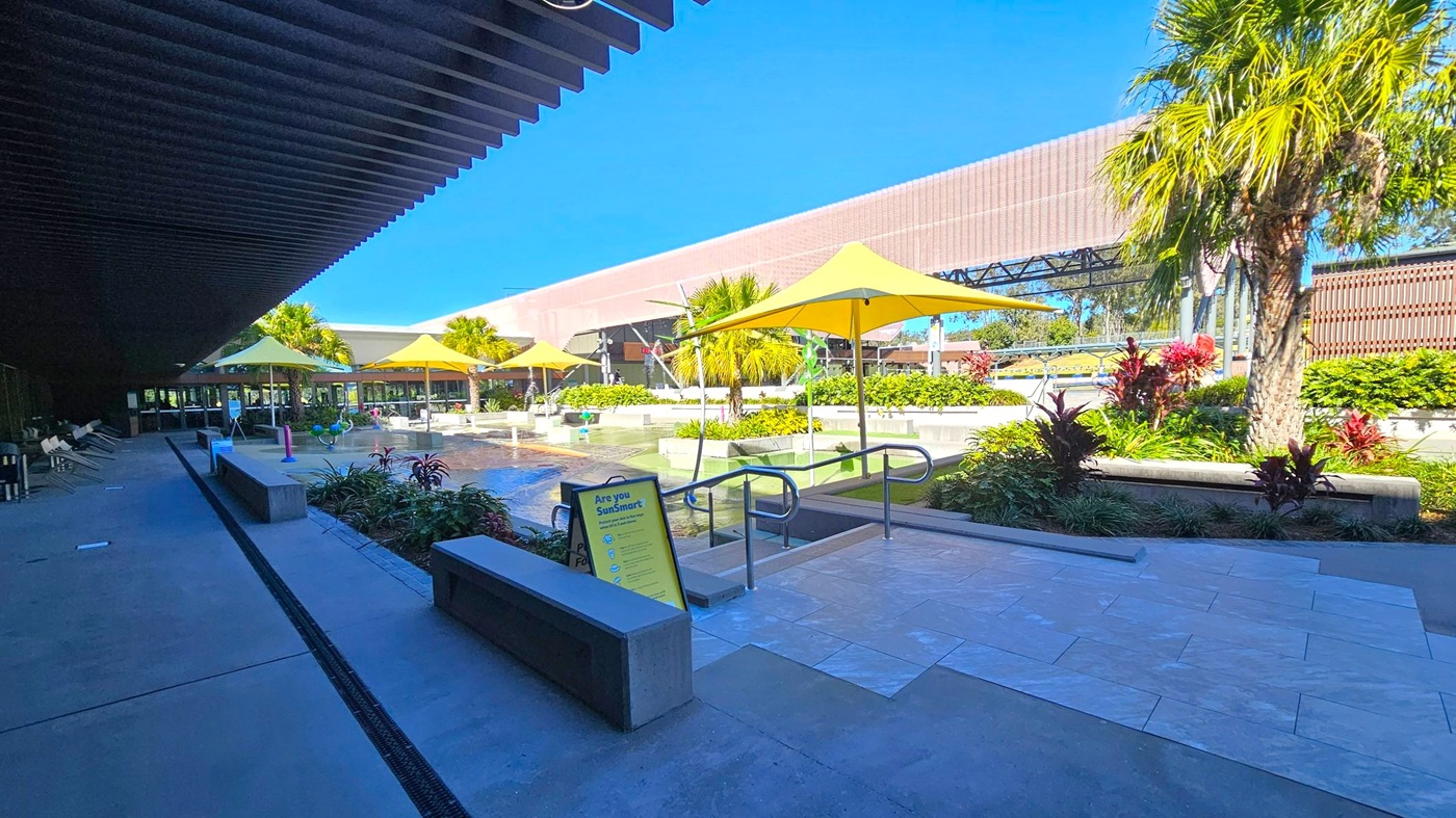 A pool area with yellow umbrellas and palm trees. On the left, there is a shaded walkway with lounge chairs and benches. In the centre, a sign reads "Are you SunSmart?" near a pool entrance with handrails. The background features a large building with a modern design and more greenery. The floor is a mix of concrete and tiled surfaces.