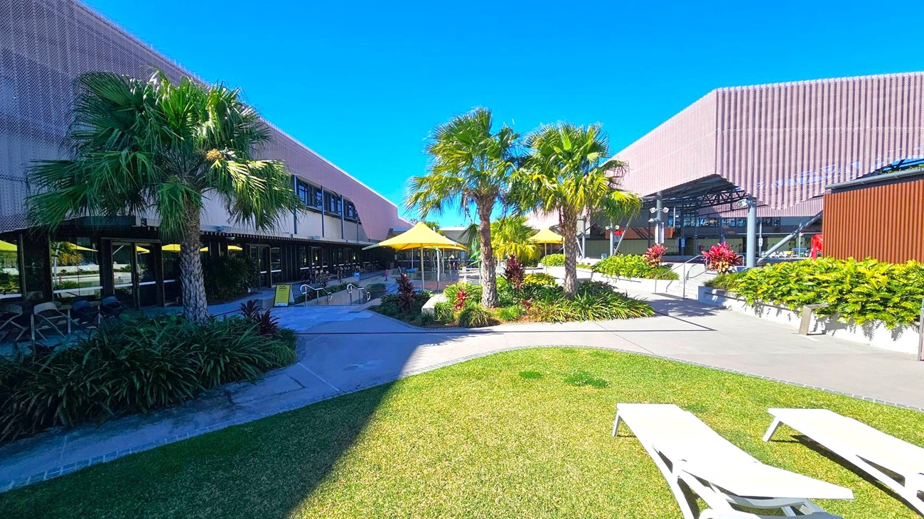 A courtyard with a grassy area in the foreground, featuring two white sun loungers. In the centre, there are several palm trees and yellow umbrellas providing shade over seating areas. The left side has a building with large windows and a covered walkway. On the right, another building with a modern design and a row of plants along a concrete path. The sky is clear and blue.