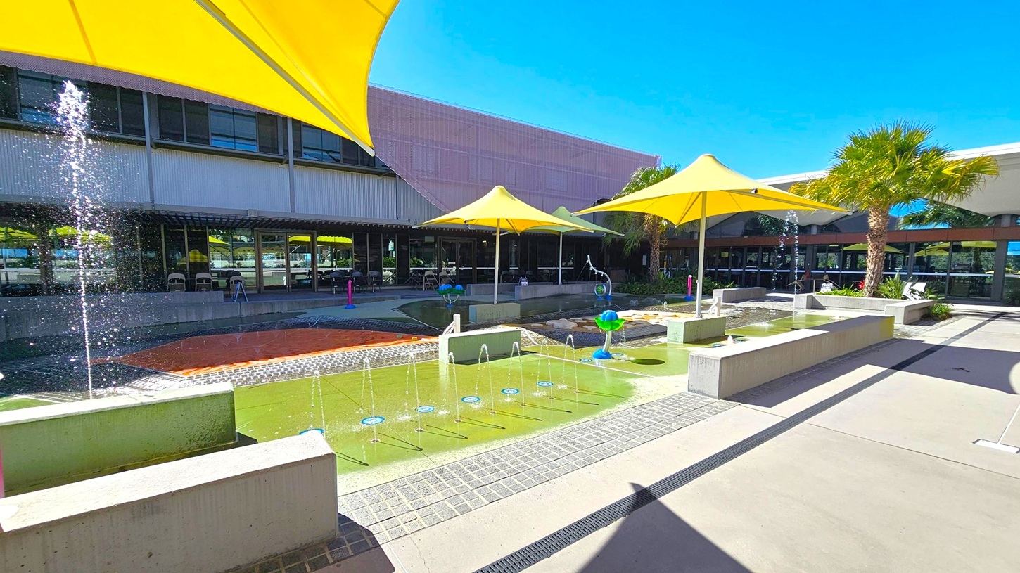 Outdoor area with a water play feature in the foreground, including fountains and a shallow pool. Yellow triangular shade structures are positioned above, providing cover. Concrete benches surround the water area. In the background, a building with large windows and a corrugated metal facade is visible. Palm trees are scattered around, adding greenery to the scene. The ground is paved with concrete, and the sky is clear.