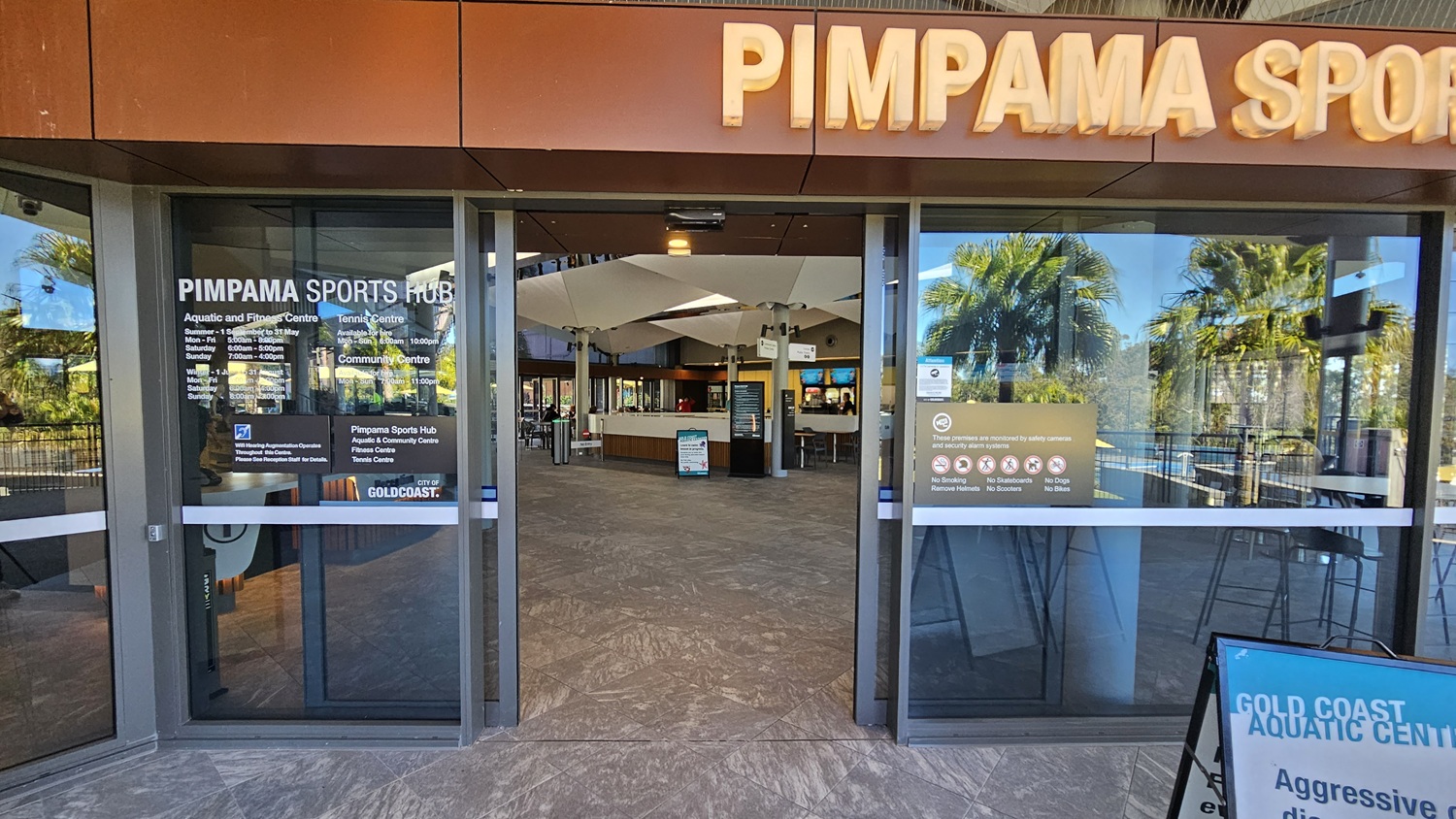 Entrance to the Pimpama Sports Hub with large glass doors. The left door displays opening hours for the Aquatic and Fitness Centre, Tennis Centre, and Community Centre. The right door has signs prohibiting smoking, skateboards, scooters, dogs, and bikes. Inside, a reception area with a counter and seating is visible. Reflections of palm trees are seen on the glass. The floor is tiled, and the ceiling has a modern design with lighting. A sign for the Gold Coast Aquatic Centre is in the foreground.