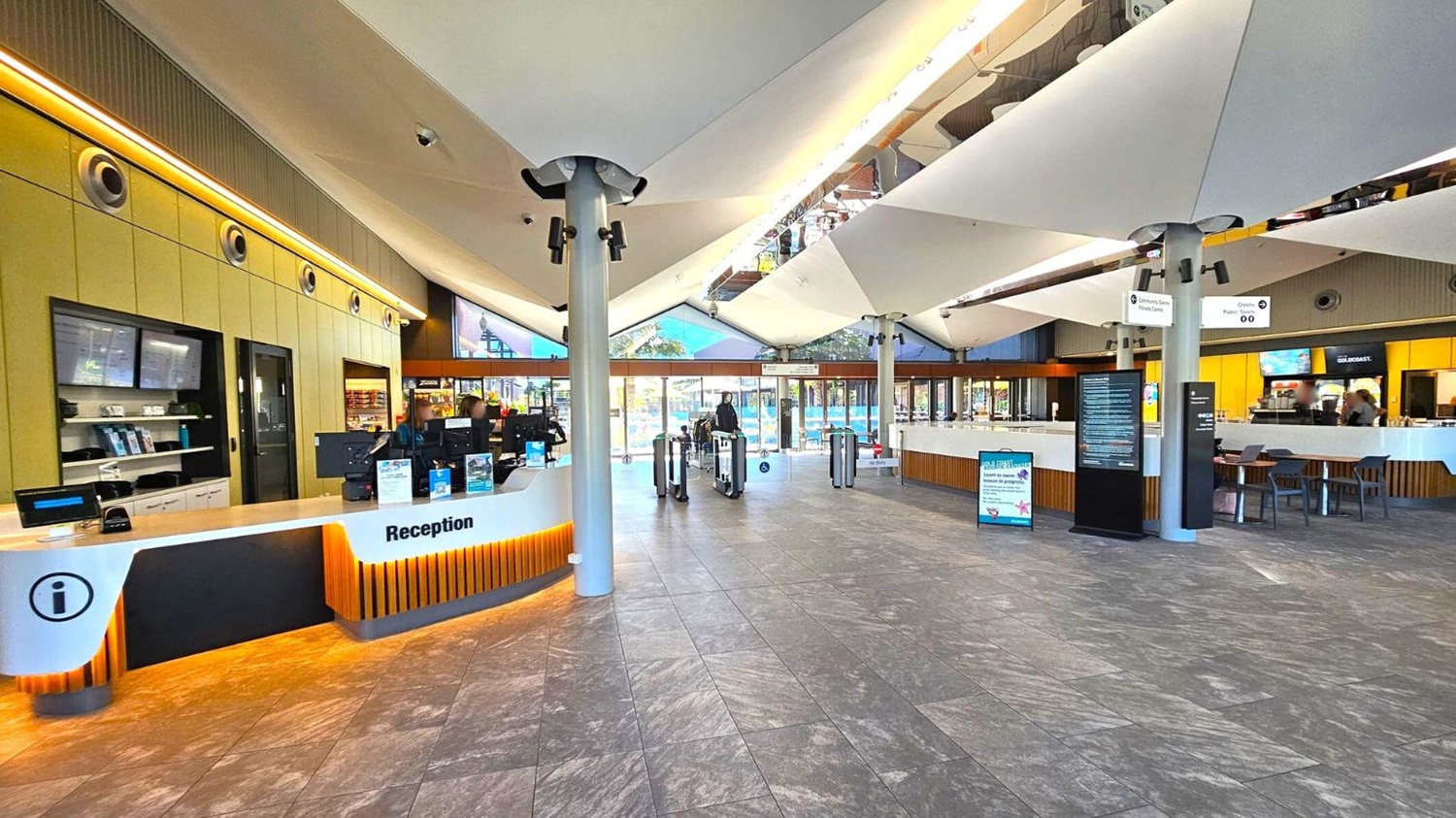 A spacious indoor area with a reception desk on the left, featuring computers and informational displays. The floor is covered with grey tiles. In the centre, there are turnstiles leading to a glass entrance with views of an outdoor area. The ceiling has a modern design with large white panels and exposed beams. On the right, there is a café area with tables and chairs. The walls have a mix of wood and glass, and there are digital screens and signs throughout.