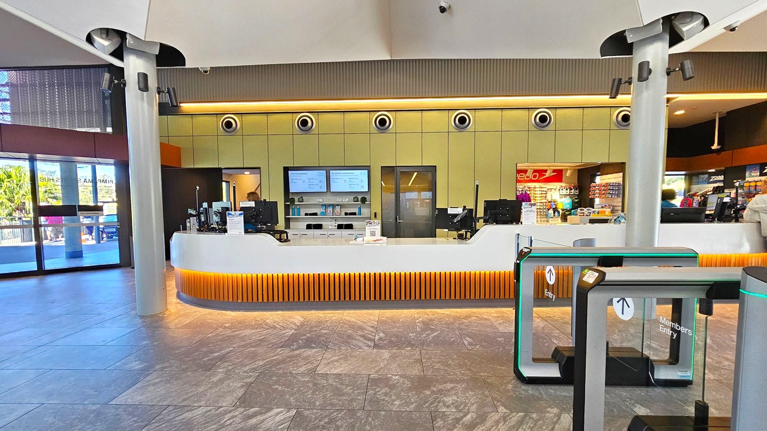 A modern reception area with a sleek, white counter in the centre, featuring orange lighting underneath. Behind the counter are two digital screens and several computers. To the right, there is a shop area with various products on display. On the left, large glass doors lead outside, allowing natural light to enter. The floor is tiled with a grey pattern, and the ceiling is white with circular light fixtures. Two tall pillars are positioned at the front.