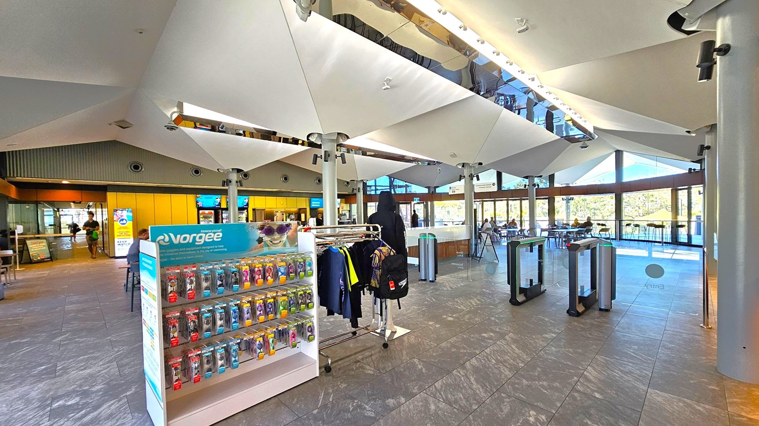 A spacious indoor area with a modern design. On the left, a display stand with swimming goggles and accessories is visible. In the centre, a clothing rack holds various garments. The floor is tiled with a grey stone pattern. The ceiling features large, angular white panels with lighting. On the right, glass entry gates lead to a seating area with tables and chairs near large windows, offering a view of greenery outside.
