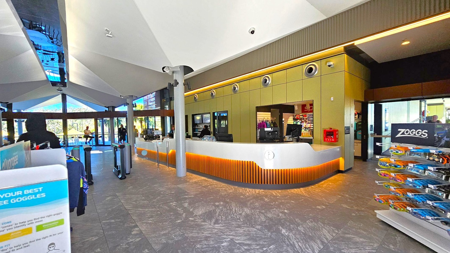 A modern interior with a reception desk in the centre, featuring a sleek white counter with wooden accents and a digital display. Behind the desk, there are shelves with various items. To the right, a display stand with swimming goggles is visible. The floor is tiled with a grey pattern, and the ceiling has a geometric design with recessed lighting. Large glass windows in the background allow natural light to enter, and a few people are visible near the entrance.