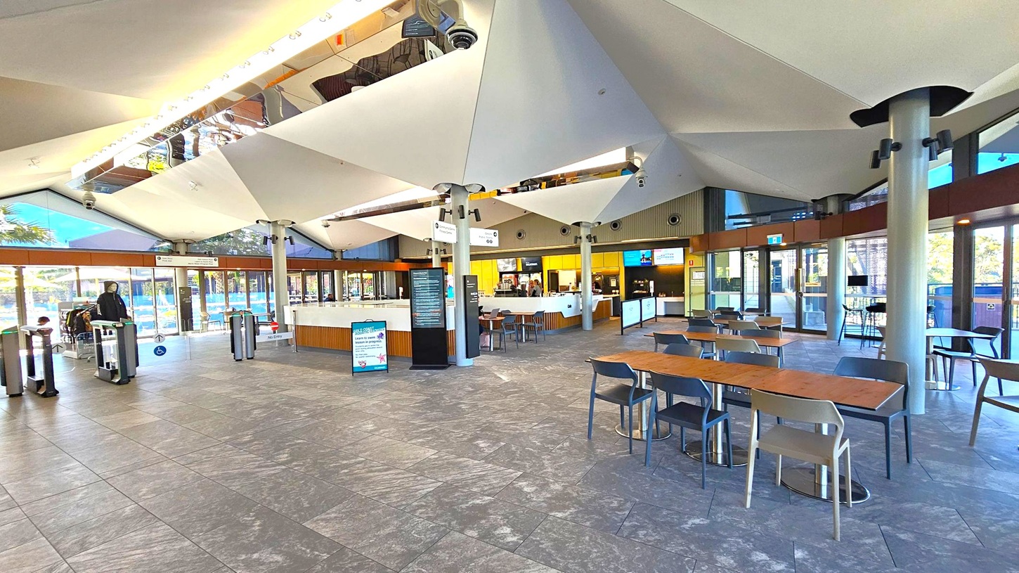 A spacious indoor area with a high, geometric ceiling featuring large white panels. On the left, there are turnstiles and a person entering. In the centre, a counter with digital screens and a menu board. To the right, several tables and chairs are arranged on a grey tiled floor. The walls are mostly glass, allowing natural light to illuminate the space.
