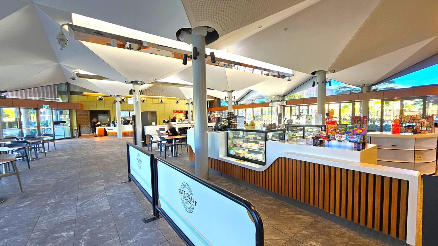 A spacious café interior with a modern design. On the right, a counter displays various snacks and a glass case with food items. In the middle, a person sits at a table with a laptop. The left side has more seating with tables and chairs. The ceiling features geometric, white panels supported by columns. Large windows in the background allow natural light to illuminate the space. The floor is tiled with a grey and purple pattern.