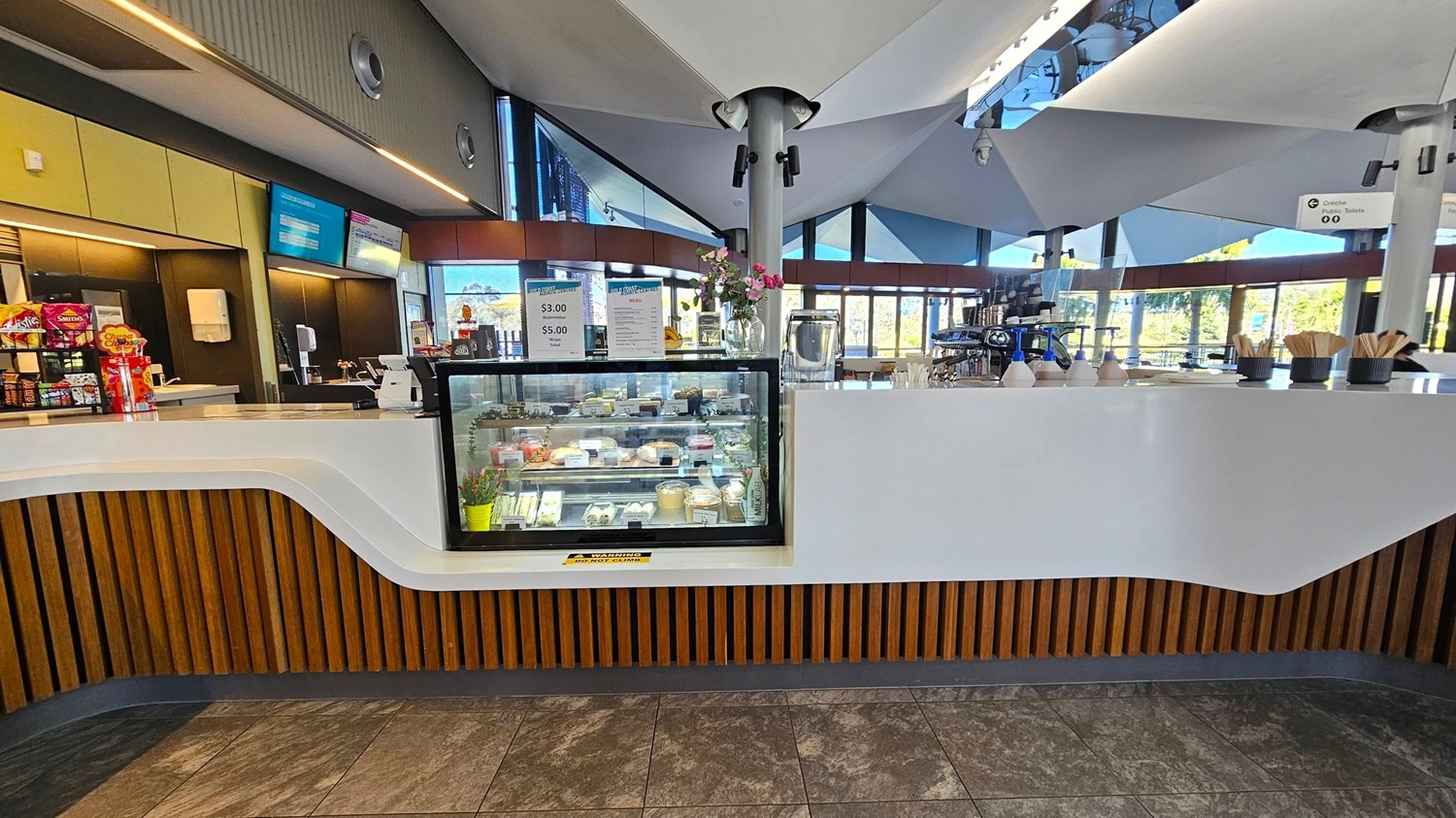 A modern café interior with a white counter featuring a wooden slat design at the base. In the centre, a glass display case contains various pastries and desserts. Above the counter, a menu is displayed on digital screens. To the right, a coffee machine and condiments are visible. The ceiling has a geometric design with spotlights, and large windows in the background allow natural light to enter. The floor is tiled with a stone-like pattern.