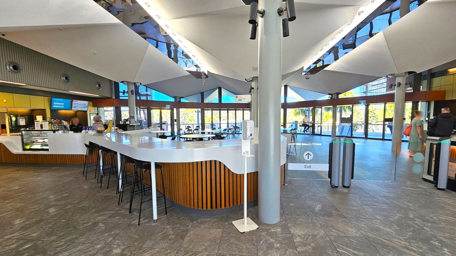 A modern indoor café area with a sleek, white counter in the centre, surrounded by high black stools. The counter has a wooden panel design and a glass display case with food items. Two people are at the counter, and a few others are scattered around the space. The ceiling features large, angular white panels with spotlights. The floor is a mix of grey and green tiles. Large glass doors in the background lead to an outdoor area with visible greenery. A hand sanitiser station is near the counter.